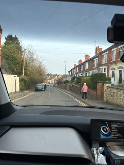 The image depicts a residential street in Wellingborough, United Kingdom, viewed from the interior of a vehicle. The foreground shows the vehicle's dashboard, with a light grey trim panel and a central infotainment screen displaying "16:39" and menu options such as "Radio," "Multimedia," and "Navigation."
The street, sloping gently downhill, has a double yellow line on the left and a dashed white line in its center. On the right, a row of red brick terraced houses with bay windows and chimneys lines the road, fronted by low brick walls and hedges. An individual with white hair, wearing a pink top and dark trousers, walks away from the viewer on the sidewalk adjacent to these houses.
On the left side of the road, a dark-colored hatchback car is parked, facing downhill. A person in dark clothing stands near the driver's side of this vehicle, appearing to handle green bags. Further along the left, there is a lighter-colored wall and dense deciduous vegetation. Overhead power lines traverse the sky, which is overcast. A street light pole is visible further down the road on the left.