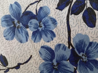 This is a close-up, overhead shot of a floral pattern on fabric, featuring blue flowers with dark centers and dark green leaves on a light cream-colored background with faint, wispy grey lines. The fabric appears to be textured. The image does not depict any specific location in Hlynytsi, Ukraine, or any discernible urban elements.