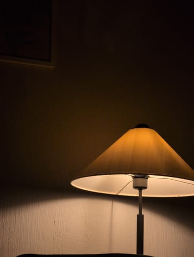 This media file features a close-up view of a wall lamp casting light on a textured wall. The lamp shade is a warm, pale yellow color and has a conical shape. The light emanating from the lamp is bright and creates a strong contrast between illuminated and shadowed areas on the wall. A dark, thin arm extends from the lamp, attached to a mounting on the wall. To the left of the lamp, a portion of a framed picture or artwork is visible against a dark background. The overall impression is one of a dimly lit indoor space, possibly at night or in a room with limited natural light. The texture of the wall suggests it might be stucco or plaster. No people are visible in the frame. The scene evokes a sense of quiet or solitude due to the limited light and absence of activity. The location context is Słupsk, Poland.