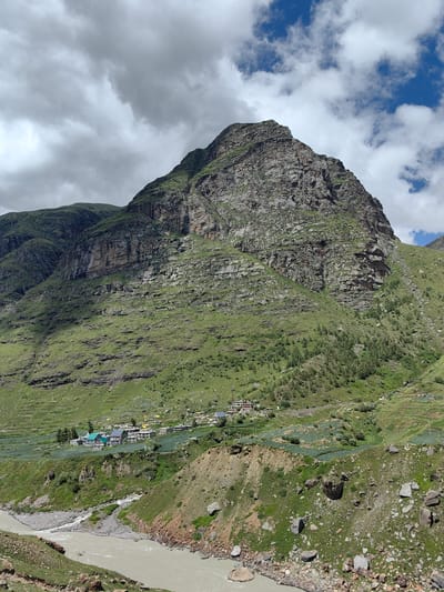 This image depicts a vast mountain landscape with a river in the foreground, likely in the vicinity of Yari-Khoksar, India. A wide, murky river flows through a rocky, sparsely vegetated riverbed, bordered by steep, green slopes. The slopes are covered with grass and dotted with trees and shrubs, indicating a rural or mountainous environment.

Higher up the mountainside, a small cluster of buildings forms a village. The structures are varied in color and style, with some appearing to be houses and others possibly more public buildings. Some of the buildings have brightly colored roofs. These buildings are situated on terraced land, suggesting agricultural activity. The terraces are covered in a green crop, possibly fields of vegetables or grains.

The dominant feature of the image is the towering mountain that rises majestically behind the village. The mountain face is rugged and rocky, with distinct layers and formations. Patches of green vegetation cling to the lower slopes, becoming sparser higher up. The sky above is partly cloudy, with a mix of bright white clouds and darker, more ominous ones, suggesting dynamic weather conditions. The overall impression is one of a serene, yet powerful, natural environment, with human settlements nestled within the grandeur of the mountains.