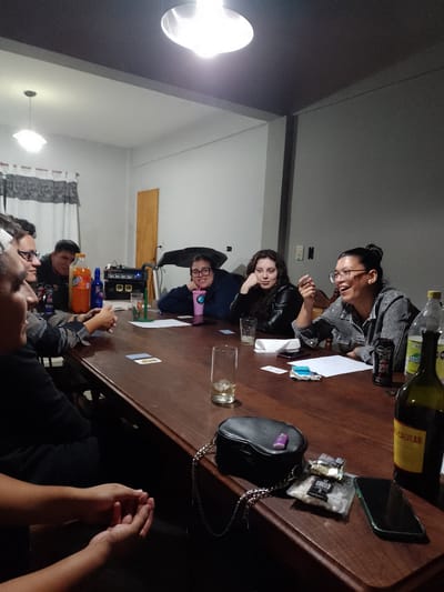 A group of seven people are gathered around a large, dark wooden table in a room with white and gray walls. The room is illuminated by two overhead lights, one round and brighter, and another square and dimmer. On the white wall is a window with curtains, and a wooden door. On the gray wall are two electrical outlets.
The people are interacting, some gesturing and talking, others listening. The table holds various items: drinks including soda and a bottle of wine (labelled Realvear), glasses, a black purse with a chain, a package of Torguitas cookies, a lighter, a smartphone, napkins, cards, and papers. The presence of these items suggests a casual social gathering.
Based on the location of the Realvear wine brand, it can be inferred that the location is Gobernador Julio A. Costa, Argentina.