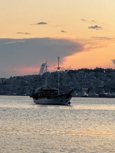 The image captures a serene harbor scene at sunset. The sky is a blend of warm orange and soft pink hues, with scattered dark clouds adding depth. A city with densely packed buildings rises up from the coastline, its lights beginning to twinkle as dusk settles in. In the foreground, a partially submerged ship rests in the water, its hull dark and weathered. A tall mast with a white banner attached stands prominently from the vessel. The water is calm, reflecting the soft light of the sky and the distant cityscape. The overall mood is one of peaceful contemplation, with the fading light and the still water contributing to a tranquil atmosphere. The scene is likely set in a port or coastal town in Greece, given the location context of Dafni. No discernible people are visible in the image. The visible text on the banner is illegible due to its distance and the image resolution.