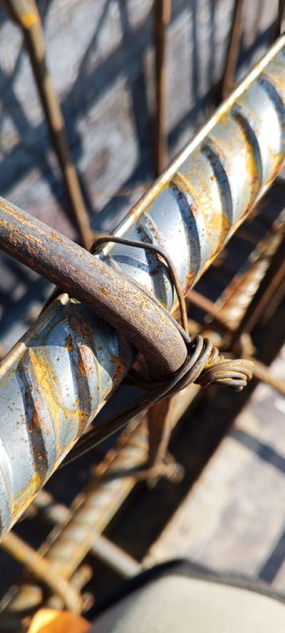 A close-up, outdoor shot shows two pieces of rebar, bound together with wire. The rebar in the foreground is rusty and dull brown, while the rebar behind it is a metallic silver color with brown rust streaks and indentations. The wire is dark and twisted, securing the two rebar pieces. The background is out of focus, showing a blurry pattern of dark lines and light shadows, suggestive of construction materials or scaffolding. The scene is illuminated by bright sunlight, creating sharp contrasts and highlights on the metallic surfaces. The image appears to be taken in an outdoor construction area.