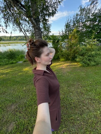 A woman with brown hair pulled back in a bun, wearing a dark brown knitted dress, stands in a grassy area next to a body of water. She is looking to her right with her left arm raised, touching her head. Behind her, a large tree with white bark stands prominently, its branches reaching towards the sky. The background features a calm body of water bordered by lush green vegetation and a distant treeline. The sky above is blue with scattered clouds. The location appears to be Burdyata, Russia, given the context.