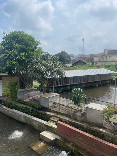 The scene captures an outdoor environment, possibly in Sumberdalem, Indonesia, based on the OCR text "TUMBU", which could be a fragment of the town's name. The viewpoint seems to be from an elevated position, looking down onto a waterway or canal. On the right side of the image, there is a waterway with evidence of water movement or a small fountain. To the left of the water, there are concrete retaining walls or ledges. On the opposite bank, a series of wire-mesh cages or enclosures are visible, possibly for keeping birds or small animals.

Vegetation is abundant, with trees and greenery spread throughout the scene. The buildings and structures in the background suggest an urban or semi-urban setting. A communication tower is also visible in the distance, adding to the context of a developed area.