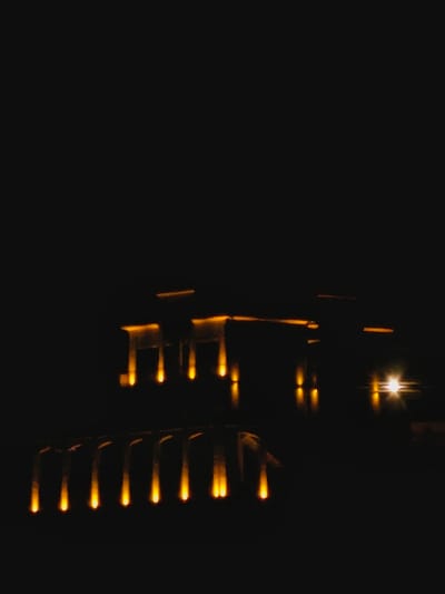 In the darkness, faint yellow light emanates from two obscure structures. The structure on the right appears like stacked barrels, each glowing along its top edge. To the left, a more angular shape flickers, also defined by yellow light. An intense pinprick of light sits above and between both. The scene is almost entirely dark, only defined by the scattered light outlining the unseen forms.