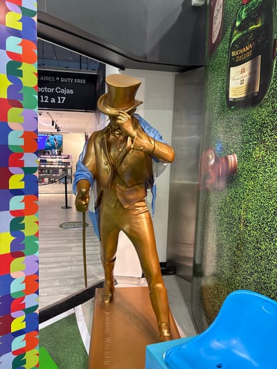 The image depicts a life-size golden statue of a man dressed in a top hat, suit, and boots, holding a cane. The statue is positioned in what appears to be a duty-free shop, possibly within an airport in Ezeiza, Argentina, given the "AIRES DUTY FREE" signage visible in the background. The statue is draped with a blue and white sash, resembling the Argentine flag. Behind the statue, a large advertisement for Buchanan's Deluxe 12 Year Old Blended Scotch Whisky is displayed on a textured green background. To the left of the statue, a colorful, abstract mural runs vertically along a pillar. A blue plastic seat is visible in the foreground on the right. The floor is tiled with a light wood pattern, and a green carpeted area is visible near the base of the statue. The overall lighting suggests an indoor commercial environment.