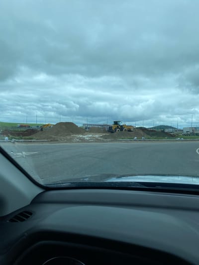 The image captures a construction site under a cloudy sky. In the foreground, the dashboard and windshield of a vehicle are visible. The construction site features large piles of sand and dirt, with a yellow front-end loader operating on one of the mounds. Several trucks, including a white semi-trailer truck and an orange dump truck, are parked or driving in the background. Streetlights are also present along a road. The scene appears to be an open area with construction activity, suggesting it could be part of infrastructure development or a new building project. The location is identified as Kliuchiki, Russia.