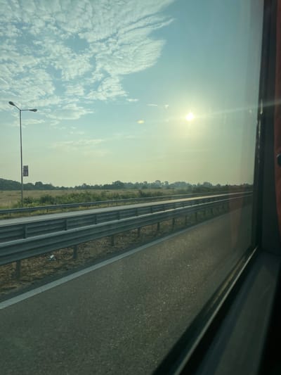 The image depicts an exterior view from the interior of a vehicle, looking out onto a highway scene in Trilistnik, Bulgaria, during daylight hours, likely morning or late afternoon given the sun's position. The sky is partially cloudy with scattered cumulus clouds against a pale blue backdrop. The sun, a bright white orb, is prominently positioned in the upper right quadrant, generating significant lens flare across the upper right portion of the frame.
A multi-lane asphalt highway occupies the lower half of the image, extending into the distance. A solid white line marks the immediate edge of the visible lane on the left. Parallel to the roadway, a two-tiered metal guardrail system is prominent, separating the asphalt from a strip of dry, light-brown vegetation and soil. A second, similar guardrail is visible further across a median, indicating a divided highway.
Beyond the guardrails and median, a flat, open field or pastureland with sparse, dry vegetation extends to a distant treeline of deciduous trees. Closer to the road, green shrubs and bushes are interspersed with the dry ground cover.
On the far left, a tall, dark grey street lamp post stands, featuring two horizontal lamp fixtures at its apex. A rectangular sign with red and blue sections, bearing white text, is affixed to the lower part of this lamp post. The right side of the image shows the dark framing and reflections of the vehicle's window. No people or animals are visible.