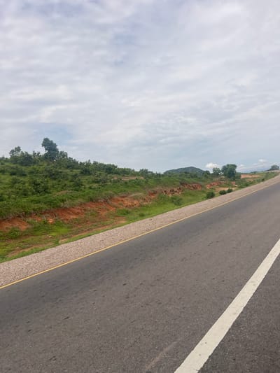 The image shows a road in Minda, Nigeria. The road is paved and has a white line marking the lane. On the left side of the road, there is a grassy embankment with some trees and shrubs. In the background, there are hills covered in vegetation. The sky is cloudy, suggesting it might be a partly cloudy day. The scene appears to be a rural or semi-rural area, given the natural landscape. There are no people or specific objects other than the road and natural elements visible in the image. The perspective suggests the photo was taken from a moving vehicle or from a vantage point beside the road. The time of day is not precisely determinable but appears to be daytime.