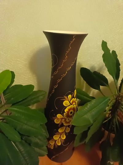 A dark, tall, slender vase with a flared rim stands on a surface, possibly a table. The vase is adorned with a swirling pattern and painted yellow flowers with red centers. It is partially obscured by lush green foliage from two potted plants, one on each side, which frame the vase. The background is a textured, pale yellow wall. The lighting appears to be artificial, casting a shadow of the vase on the wall to the left. The image is taken from a slightly low angle.