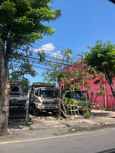 This is a vertical, eye-level shot of a street scene in Denpasar Barat, Indonesia, under a clear blue sky. The image is dominated by two large trucks parked side-by-side along a busy road. The truck in the foreground is white with black accents, and it has the word "Bismillah" written in white script on its side. The second truck, parked behind it, is green. Both trucks appear to be parked on the side of the road, near a concrete curb.
Trees with lush green foliage and exposed branches partially obscure the trucks and a bright red building behind them. The sidewalk is littered with debris, including plastic bags, leaves, and other unidentifiable items, suggesting a somewhat neglected urban environment. Electrical wires are strung overhead, adding to the urban feel. The overall impression is one of a sunny day in a tropical city, with the trucks being the primary focus, possibly indicating a commercial or transportation hub. The time of day appears to be mid-morning or early afternoon, given the bright sunlight and distinct shadows. There are no people visible in the image.
Visible text includes: "Bismillah" on the white truck, and "www.karyausahattran.co." on a sticker on the windshield of the truck behind the white one. License plates are also visible on both trucks, with the white truck's plate reading "AG 1388 US" and the lower truck's plate reading "AC 8898 UU".