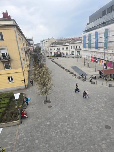 An elevated view captures a pedestrianized urban street in Nyíregyháza, Hungary, specifically the area adjacent to the Korzó Shopping Center, under an overcast sky. The ground is paved with grey interlocking blocks.
To the left, a multi-story light yellow building with numerous windows and balconies runs parallel to the street. A narrow landscaped strip with low green hedges and two small deciduous trees with white blossoms borders this building. A street lamp with hanging flower baskets is positioned in the foreground. Two individuals are seated on a bench in this area, one wearing a red jacket. A delivery person, dressed in a blue jacket and carrying a blue "Wolt" delivery bag, stands next to a bicycle near the bench. Another individual walks further along the sidewalk.
The central paved area features multiple people walking. Two uniformed individuals, wearing dark blue clothing with yellow reflective vests, walk side-by-side. Behind them, two other individuals, including one with visible reddish hair, walk together. Scattered across the pedestrian zone are various dark square planters and several sets of outdoor tables and chairs. A dark metal strip is embedded in a section of the paving.
To the right, the modern Korzó Shopping Center dominates the scene with a stone-clad and glass facade. Four vertical banners displaying "KORZÓ" in white letters, alternating teal and blue, are affixed to the building. Large commercial advertisements featuring faces and text, including "HEAVY TOOLS," are visible on its lower section. A small, dark-roofed kiosk labeled "Kürtőskalács" is situated on the paved area in front of the shopping center, with additional outdoor tables and chairs.
Distant urban buildings of various architectural styles are visible in the background, receding under the uniform grey sky.
