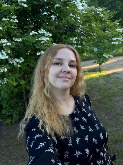 A young woman with long, wavy, light blonde hair is depicted in a close-up outdoor portrait, situated in Urmanets, Russia. She faces directly towards the camera, her head slightly tilted to her right, and exhibits a soft smile. Her eyes are light-colored, and her fair complexion displays visible freckles. She is wearing a black dress adorned with a repeating pattern of small, stylized white floral or foliage motifs.

The background on the left side of the frame features dense green foliage from a flowering shrub, characterized by numerous clusters of small white blossoms. To the right of the woman, a light brown dirt or gravel path is partially visible, illuminated by warm, direct sunlight that creates distinct shadows. Patches of green grass and sparse low vegetation line the path. The overall lighting suggests late afternoon or early evening conditions.
