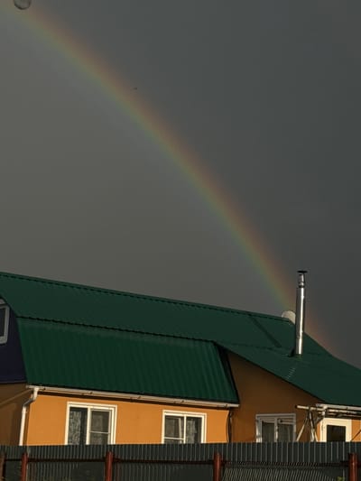A bright rainbow arcs across a grey, overcast sky, its vibrant colors a stark contrast to the muted atmosphere. The rainbow's arc begins on the left side of the frame and extends towards the right, partially obscured by the roof of a building. The building itself is a striking feature, with a green corrugated metal roof angled sharply downwards from the right edge of the image. Below the green roof, an orange facade is visible, featuring several white-framed windows. One window near the top of the orange section has blinds. Another window below it is partially covered by what appears to be patterned fabric or wallpaper. A third window is visible at the bottom of the orange section. A metal pipe, possibly a chimney or vent, extends horizontally from the green roof. On the far right of the image, a dark grey corrugated metal structure runs vertically, with horizontal wooden beams attached at intervals. The overall scene suggests a residential area under a cloudy sky, possibly after a rain shower, given the presence of the rainbow. The lighting indicates it is daytime. The location context provided is "Южное Снт, Russia," which helps to place the scene within a specific geographical area. There is no visible text in the image.