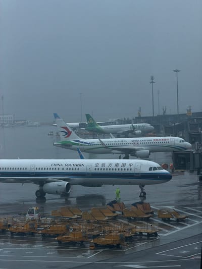 The image depicts an airport tarmac on a misty or rainy day, with several airplanes parked and a ground crew member visible. In the foreground, the front section of a China Southern Airlines airplane is prominent, showing its fuselage with "CHINA SOUTHERN" written in English and Chinese characters. The airplane's landing gear is visible, and the tarmac is wet, suggesting recent precipitation.

Behind the China Southern plane, a China Eastern Airlines aircraft is partially visible, also with its airline name clearly marked. Further back, another airplane with a green tail and a stylized 'S' logo, likely belonging to Sichuan Airlines, is present. The planes are positioned near jet bridges or terminal structures.

Numerous yellow luggage carts are parked in rows on the tarmac, some laden with cargo. A small utility vehicle is also visible near the carts. A single individual, wearing a bright yellow fluorescent jacket, is walking on the tarmac in front of the China Southern Airlines plane, appearing to be a ground crew member. The overall atmosphere is subdued due to the weather conditions, with the mist obscuring the background details and the sky. The location is an airport in Guangyang District, China.