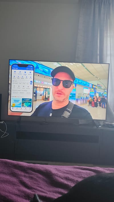 A television screen displays a video of a man wearing sunglasses and a cap, with a phone overlay showing the Trip.com app. The man is standing in what appears to be an airport. The background shows airport terminals with various signs and people. The phone screen displays travel options such as flights, hotels, and train tickets. The overall scene is illuminated by artificial lighting, consistent with an indoor environment. The television is positioned on a dark media console. In the foreground, a plush purple blanket is visible, partially covering a dark object, potentially furniture or a pet. The image is taken from a low angle, suggesting the viewer is seated. The location is identified as Nemėžis, Lithuania, but this information is not visually ascertainable from the image itself.