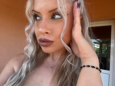 A woman with blonde hair and dramatic makeup is posing for a selfie outdoors. She is wearing a black and gold bracelet. The background appears to be a residential area, possibly a courtyard or garden, with an orange wall and a glass door. The lighting suggests it is daytime. The woman's pose and expression suggest confidence and allure.