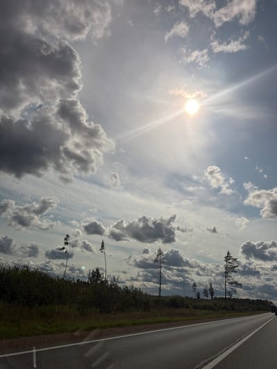 The image shows a road in Odukalns, Latvia, with a dense green forest on the left. The sky is partly cloudy with the sun visible and emitting bright rays. There are a few trees scattered throughout the landscape. The road is asphalt and has white lane markings. The scene is captured from a low angle, suggesting it was taken from a vehicle.