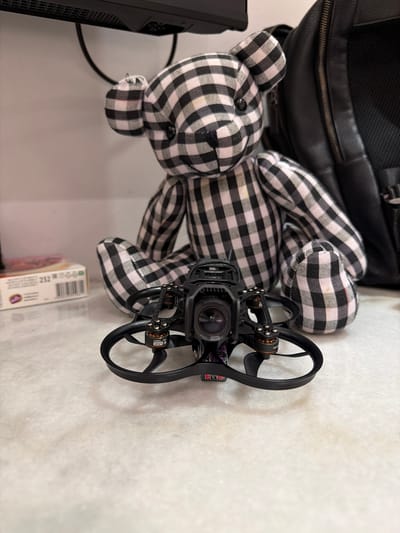A miniature drone with black propellers and orange motor accents sits on a white marble countertop. Behind the drone, a plush teddy bear with a black and white checkered pattern is seated. To the left of the bear, a box with text and a barcode is partially visible. To the right of the bear, a black leather backpack is visible. The background is a plain white wall, and a black monitor is perched at the top of the frame. The scene is indoors, suggesting a home or office environment. This image is not located in Ban Phonsavang Neua, Laos.