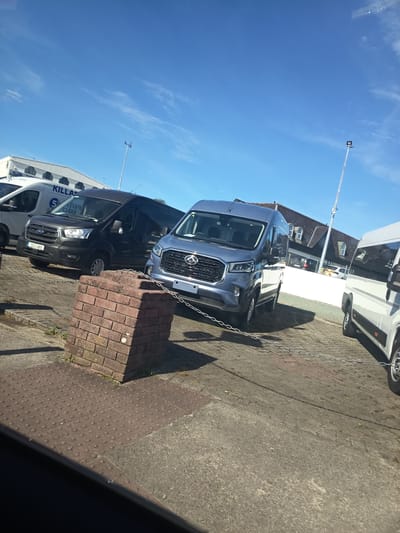 The image shows a parking area in Killarney, Ireland, with several vans parked on a paved surface. The sky is blue with some scattered clouds. In the foreground, a brick pillar is visible, along with a chain link barrier. Several vans are parked in the background, including a black Ford Transit van with a license plate starting with "GC". A light blue van is parked to the right of the black van, and a white van is partially visible on the far right. A white building with arched windows is visible behind the vans. A tall light pole with a lamp is also present.