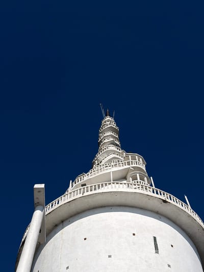 A low-angle shot reveals a tall, white tower against a deep blue sky. The tower's architecture features a cylindrical base with a spiraling, cage-like structure that ascends and narrows towards the top. Ladders and walkways encircle the tower, adding a sense of height and complexity. The perspective emphasizes the tower's upward reach, contrasting its bright, man-made structure with the vastness of the sky.