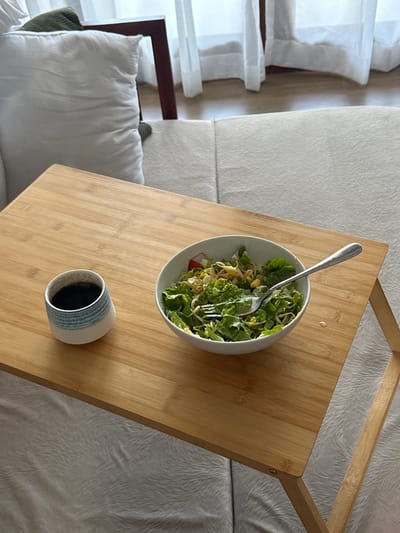 In a bright indoor setting, likely a living room or bedroom with natural light filtering through sheer curtains, a wooden tray table is placed on a textured white couch. The tray holds a small, blue and white striped ceramic cup filled with dark liquid, presumably coffee, and a white bowl overflowing with a fresh salad. A silver fork rests within the salad, indicating it is ready to be eaten. In the background, a large white cushion and a portion of a dark wooden chair are visible, suggesting a comfortable and relaxed atmosphere. The overall impression is one of a peaceful meal enjoyed indoors, with no specific indication of the city of North Kuta, Indonesia.