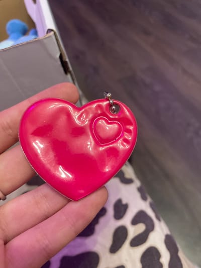 A close-up shot shows a hand holding a bright pink heart-shaped charm with a smaller embossed heart in its center. The charm has a small metal loop at the top, indicating it's likely a keychain or a pendant. The background is blurred, with hints of a cardboard box and a wooden surface, suggesting an indoor setting. There is no discernible information to place this image in Vaganovo Massiv, Russia.