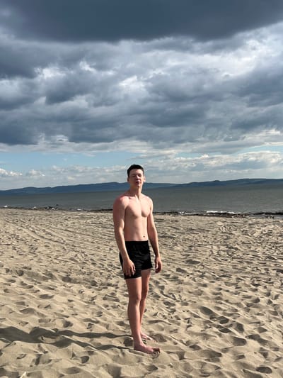 The media file shows a young man, shirtless and wearing black swim trunks, standing on a sandy beach. He is facing the camera with his arms relaxed at his sides. The beach is covered in fine, light-colored sand with distinct ripples and textures. In the background, a body of water stretches out, meeting a distant mountain range under a dramatic sky. The sky is filled with a mix of dark, brooding storm clouds on the left and brighter, lighter clouds with patches of blue sky on the right, suggesting a dynamic weather front. A few birds are scattered on the beach near the water's edge. The scene appears to be set during the daytime, given the lighting conditions and shadows cast on the sand. The overall mood is casual and serene, despite the imposing clouds. There is no visible text in the image. The location context is Krasnoturansk, Russia.