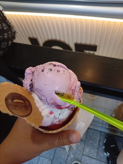 The image shows a hand holding an ice cream cone filled with two scoops of ice cream and a bright green plastic spoon. The top scoop is a light purple color with dark flecks, while the bottom scoop appears to be white with red swirls. The ice cream cone is decorated with a waffle cookie imprinted with a logo. The hand holding the cone appears to be a light skin tone. In the background, a black countertop is visible with a partially visible white wall behind it. The floor beneath the hand is a gray tiled surface. The location is Malko Chochoveni, Bulgaria.