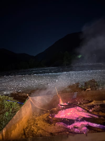 The scene depicts a nighttime environment, possibly a river valley in Sindieti, Georgia, based on the context. A dark mountain silhouette dominates the left side of the frame, partially obscured by low-lying clouds or fog. Next to the mountain, there's a wide, rocky riverbed or stream, with the water reflecting the faint ambient light.
On the right side of the frame, there's a structure, possibly a retaining wall or part of a building. Next to the structure is a burning fire. The burning wood has a visible orange glow with bright red hot spots. Smoke emanating from the fire partially obscures the surroundings. There's a mixture of soil and vegetation nearby, suggesting a natural or semi-natural setting.