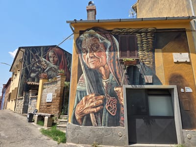 The image depicts a mural on the exterior of a building in Viterbo, Italy. The mural on the right side of the building is a close-up portrait of an elderly woman with deeply wrinkled skin and gray hair, wearing a headscarf and traditional clothing. She holds a pendant in the shape of a heart with a tree growing from its center. The building's facade is painted a warm yellow, and features a window with brown shutters and a planter box with greenery. To the left, another building displays a mural of a young woman with red hair and a wolf. Both murals are large-scale and appear to be part of an urban art project. The scene is set on a sloped street with a cobblestone surface, bordered by stone walls and a metal gate. A stone bench is situated next to the gate, with a trash can nearby. The sky is clear and blue, indicating a sunny day. The overall impression is one of vibrant street art decorating the historic urban landscape of Viterbo.