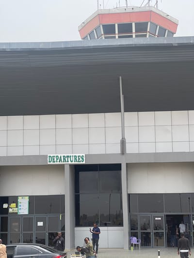 The image depicts the exterior of an airport building, specifically the departures area. A control tower is visible to the left. The building is constructed of gray ribbed metal and white rectangular tiles. A green sign with the word "DEPARTURES" is vertically aligned. The entrance consists of multiple glass doors. Several people are visible outside the entrance, along with a dark-colored car. The sky appears overcast. The location is Kwi, Nigeria.