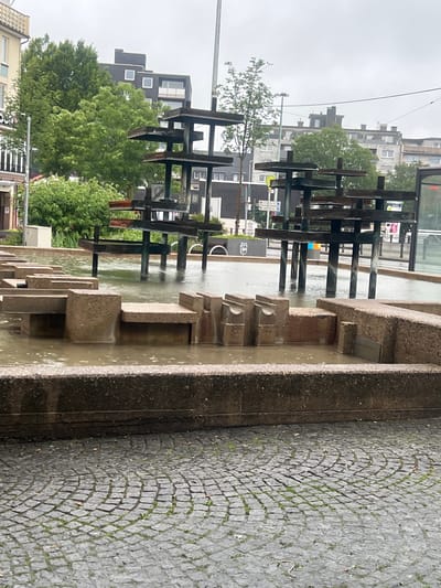 The image shows a public fountain in Bochum, Germany. The fountain is made of concrete and metal, with several tiers and water features. It is located in a paved area with cobblestones. In the background, there are buildings and trees. The sky is overcast, suggesting that it is a cloudy day. The water in the fountain appears murky. There are no people visible in the image. The overall scene is one of urban public space, likely a plaza or square.