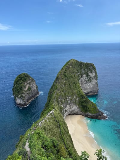 The image presents an aerial view of Nusa Penida's iconic Kelingking Beach, situated near Bunga Mekar, Indonesia.
A distinctive, T-Rex-shaped cliff dominates the frame. Its vegetation-covered spine descends steeply towards a secluded beach. The beach displays golden sand and turquoise waters. The ocean is a deep blue in the foreground.
A small, rocky island stands detached from the main cliff face. A visible pathway descends down the cliff, revealing the presence of people.