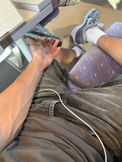 A person's arm and hand are visible in the foreground, extending towards the left side of the frame. The arm is tanned and has prominent veins. The hand is open, with fingers slightly spread. The person is wearing a grey t-shirt and dark denim shorts. A white cable, possibly a phone charger, lies across the shorts. In the background, the person's legs are visible, wearing white socks and athletic shoes. One leg rests on a seat with a purple patterned cushion. The setting appears to be on a mode of transport, possibly a train, given the presence of a tray table on the left side of the image, which has a speckled surface and is attached with hinges. The floor is a light brown color. The image does not contain any information that would allow for identification of a specific location in the city of Rivesaltes, France.