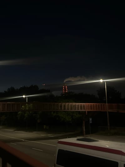 An elevated pedestrian bridge, illuminated by streetlights, spans a street at night. The bridge has a reddish-brown railing and a latticed walkway. In the background, a tall industrial tower emits smoke, with red lights visible on its structure. Trees line the area around the bridge, and a road with white lane markings is visible below. A white van with a red stripe is parked on the right side of the frame, facing away from the camera. A blue parking sign is mounted on a pole near the bridge. The sky is dark, with some wispy clouds. The overall scene suggests an industrial area or a city at night.