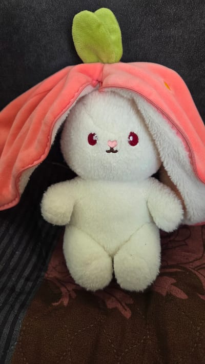 A white plush rabbit toy with red eyes and a pink nose is lying on its side, partially encased in a pink plush strawberry-shaped hood with a green stem on the left side of the frame. The rabbit's body is mostly out of the hood, revealing its fluffy white fur. The background is a mix of textures, including dark gray fabric on the left and a brown and dark brown patterned fabric on the right. The lower portion of the image contains dark gray and black striped fabric.
