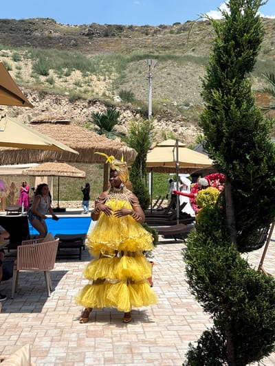 A Black woman in a vibrant yellow, layered dress and a matching headscarf, adorned with elaborate gold jewelry, stands on a brick patio. She is positioned in the foreground, facing slightly to her left, with her hands gesturing forward. Behind her, a bright blue swimming pool is visible, bordered by lounge chairs and thatched umbrellas. In the background, a steep, rocky hillside with sparse vegetation rises under a clear blue sky. To the right of the woman, a tall, manicured green tree stands prominently, partially obscuring other individuals and decorative floral arrangements in the mid-ground. To the left, other people are observed near the pool, with one woman in a pink dress and another in a white top and denim shorts. The scene appears to be an outdoor resort or event space in Ptghni, Armenia.
