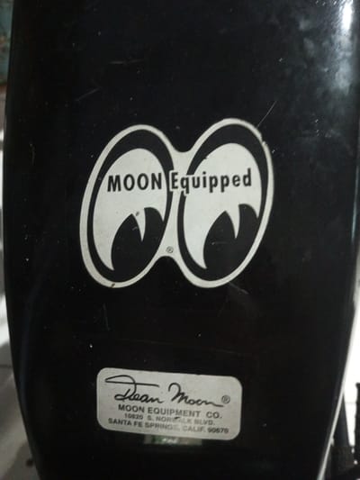 A black surface showcases two distinctive "moon eyes" logo bearing the words "MOON Equipped" above. Below the logo is a rectangular sticker with "Dean Moon" written in cursive at the top, followed by "MOON EQUIPMENT CO." and the address "10820 S. NORWALK BLVD., SANTA FE SPRINGS, CALIF. 90670" in block letters.