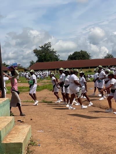 In Orun Ekiti, Nigeria, a group of individuals in white uniforms and green caps are gathered outdoors on a dirt ground. Some are walking, while others appear to be standing or in formation. In the foreground, a woman with a vibrant pink and green garment walks with a basket of water bottles balanced on her head. The background features buildings with red roofs and trees under a cloudy sky.