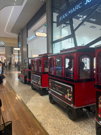 The image depicts an interior scene within a modern shopping mall, likely Cap 3000 in Saint-Laurent-du-Var, France. The primary subject is a multi-car, trackless miniature train, painted red and black, moving from left to right across the frame. The train consists of a lead locomotive and at least three passenger cars. Each passenger car features side panels with festive, Christmas-themed graphics, including candy canes and a reindeer, along with French text that reads "SANTA AMBASSADOR," "DEMON PORTEUR DE BEAUTE," and "TARIF."
Multiple individuals are visible inside the train cars, with a male adult observer with a beard identifiable in the second passenger car from the front, looking towards the right. Other passengers are less distinct due to motion blur.
The mall environment features polished light brown wooden plank flooring in the immediate foreground, transitioning to a speckled light-colored terrazzo-style floor where the train operates. Retail storefronts line the concourse; "ObeyYourBody" is prominently displayed on a lit sign above a white shop, and "JEAN MARC J" (likely Jean Marc Joubert) is visible on a large black sign above a glass facade on the right. Further into the background, "JM WESTON" and partially visible "MARC JACOBS" signs indicate additional retailers. The mall architecture includes high ceilings with linear and square artificial lighting fixtures, and large glass storefronts with dark structural framing. Several additional individuals are observed walking in the background, some carrying shopping bags, engaged in typical mall activities.