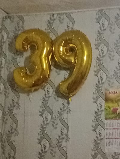 The image shows two large, gold Mylar balloons shaped like the numbers "3" and "9," forming the number "39." The balloons are inflated and appear to be hanging on a wall with patterned wallpaper. A small portion of a calendar or poster with the year "2026" printed on it is visible to the right of the balloons. The overall scene appears to be an indoor setting, likely a room. There are no people or specific landmarks visible that would indicate a location in Atbasar, Kazakhstan. The image is a close-up of the balloons and the wall.