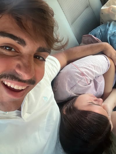 A selfie shows two people in a car. A man with brown hair and a mustache smiles broadly, taking the photo. Behind him, a woman with dark hair rests her head on him, appearing to be asleep. She wears a light purple shirt. They are both in a car. The car's interior is visible, including the gray upholstery of the seats. The woman's head is resting on what appears to be the man's shoulder.