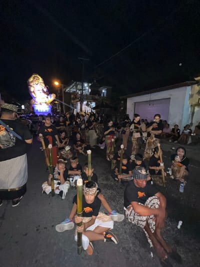This nighttime scene captures a street in Munggu, Indonesia, during a public gathering. The environment is dark, illuminated by streetlights, building lights, and a prominent, brightly lit effigy in the background.
The central subject is a large, elaborate effigy, possibly an Ogoh-Ogoh, positioned on a raised platform in the mid-ground. The effigy depicts a humanoid figure with prominent hair or a mane, seated with legs bent, and is primarily illuminated by internal blue and white lights.
A dense crowd of people occupies the paved road. Many individuals, including numerous children and adults, are seated directly on the asphalt in the foreground and mid-ground. Most are wearing black t-shirts, some featuring an orange logo. Many male participants, particularly children and men in the foreground, wear traditional Balinese sarongs (kain) and head coverings (udeng). Numerous seated individuals hold tall, slender bamboo poles, some with white tips. Actions include observing the scene, resting, and casual conversation; some individuals are engaged with mobile phones.
The street is lined with buildings. On the right, a white building with a closed metal roll-up door is visible. Further down the street on the left, multi-story buildings with balconies and illuminated interiors are present. Overhead, the dark night sky is visible, with power lines crisscrossing. Scattered small white debris and a plastic water bottle are visible on the road surface.