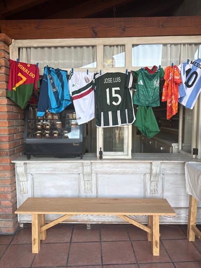 A photograph taken in Tepeji de Ocampo, Mexico, shows a collection of soccer jerseys hanging by clothespins from the frame of an open window. Starting from the top, the jerseys are: Argentina (Messi, number 10), Mexico (Puma), Mexico (green), Mexico (Jose Luis, number 15), Mexico (white), Manchester City (blue), and Portugal (Ronaldo, number 7). The jerseys appear to be drying, suggesting they have been washed. Below the window, on the exterior brick ledge, sits a small wine refrigerator filled with labeled glass jars, potentially containing local spices or condiments. To the right of the window, affixed to the wall, is a rustic wooden shelf structure with a shelf. The window frame is painted white. The photograph appears to have been taken outdoors during the day, with natural light illuminating the scene.