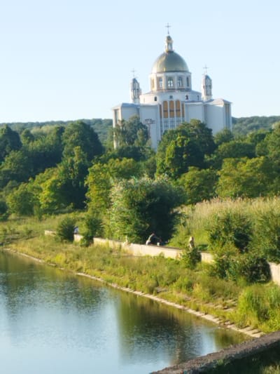 The image depicts a serene riverside scene in Novyi Rozdil, Ukraine, dominated by the striking presence of the Church of the Holy Archangel Michael. The church, a grand edifice with a prominent golden dome and white facade, stands regally against a clear blue sky. It is surrounded by lush green trees that cascade down towards the river.

In the foreground, the calm waters of a river reflect the bright sky, with a textured stone embankment lining its edge. Along this embankment, several individuals are present. Two people are seen seated, appearing to be in quiet conversation. Further along, a single person stands near the water's edge, observing the surroundings. The vegetation along the riverbank is a mix of tall grasses and shorter green shrubs, adding to the natural beauty of the setting. The overall atmosphere is one of peace and tranquility, with the architectural marvel of the church providing a focal point amidst the verdant landscape.