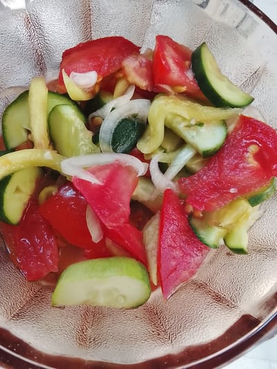 This is a close-up, top-down shot of a chopped salad in a textured glass bowl. The salad contains sliced tomatoes, cucumbers, and white onions. The ingredients are mixed together and appear to be slightly macerated, with some liquid present in the bowl. The bowl itself has a rough, pebbled texture on the inside and a dark rim. The background is blurred and appears to be a light-colored surface. The location in Kerben, Kyrgyzstan, is not discernible from the image.