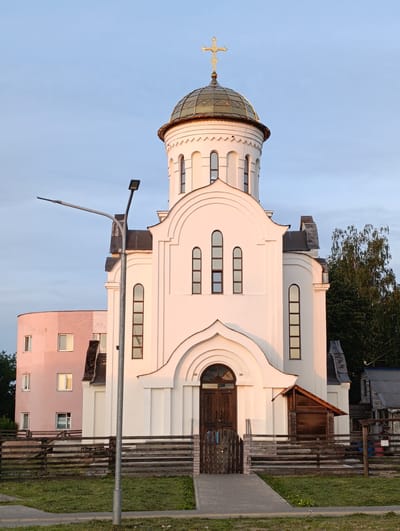 The image shows a white church with a golden dome topped with a cross. The church has arched windows and a large wooden door. In the background, there is a pink building and some trees. A lamppost stands in front of the church. The scene appears to be set during the golden hour, as the setting sun casts a warm glow on the church. The sky is a clear blue with some wispy clouds. The location is Orsha, Belarus. There are no people visible in the image. The overall mood is peaceful and serene.