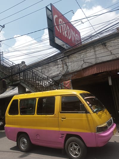 A yellow and pink minibus is parked on a street in Xayabury District, Laos. Overhead are numerous electrical wires and a large sign that reads "Cardinal." A metal staircase rises from the street to an upper level of a building. The building itself is constructed of aging concrete. The sky is partly cloudy.